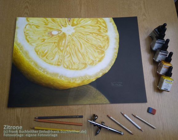 Ein gelbes Zitronenbild auf Papier, umgeben von Zeichenmaterialien und Airbrushfarben.