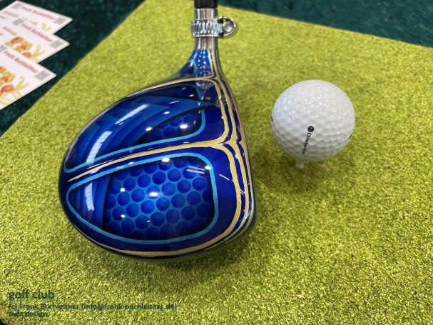 Blauer Golfschläger mit detailliertem Muster, daneben ein weißer Golfball.